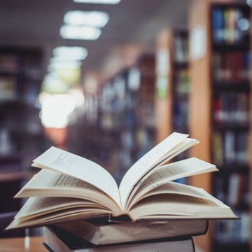 Open book on a stack in a library with bookshelves blurred in background.