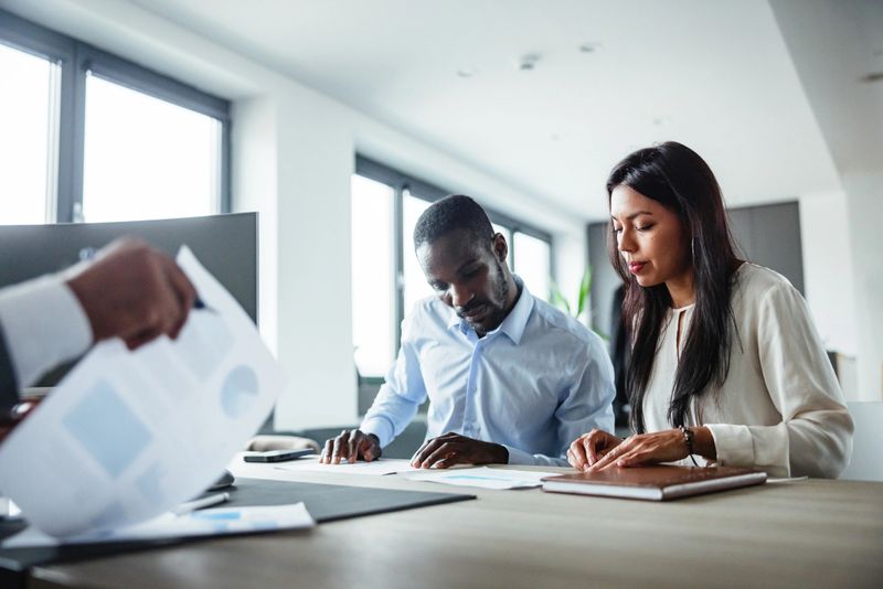 A Black man and a Latin woman engage in a business meeting, focusing on documents at a modern office desk. Their collaboration signifies teamwork and professionalism in a corporate environment filled with natural light.