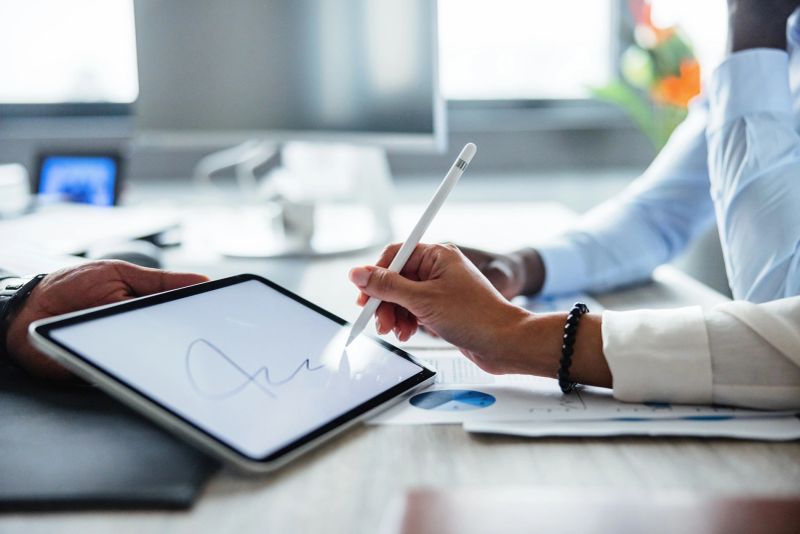 Close-up of diverse business professionals signing a digital document on a tablet in a modern office setting. The focus is on hands using a stylus, highlighting technology, collaboration, and digital contracts in corporate environments.