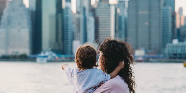 A woman holds a child pointing towards a city skyline across the water.