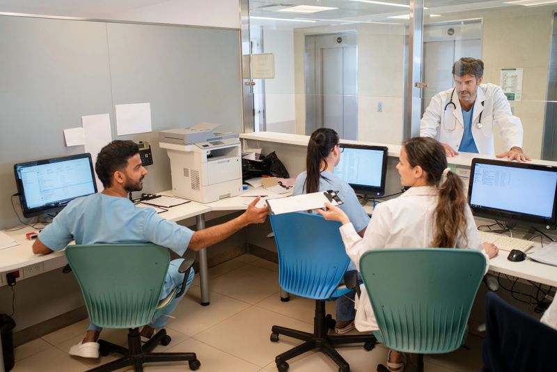 Hospital front desk staff engaged in their tasks, with a doctor standing in the office. Administrative staff woking at hospital office with doctor standing at front desk.