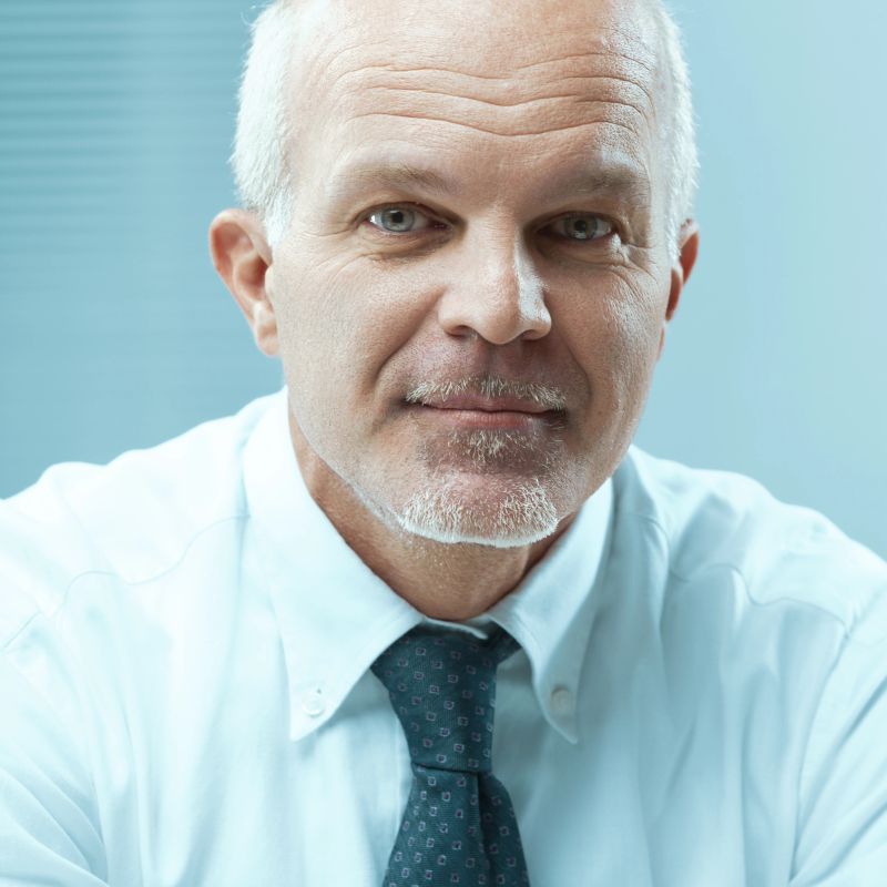 Mature businessman with a salt and pepper beard wearing a shirt and tie looking at the camera with a confident expression