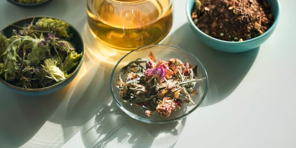 Bowls of dried herbs and a glass teapot with herbal tea on a white surface.