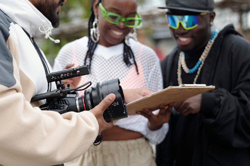 Filming crew holding camera and reviewing script on clipboard during an outdoor film shoot. Team members focused on scene planning without direct eye contact with camera