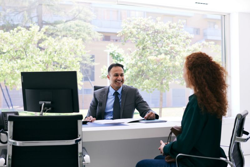 Sitting front to front at the office desk, the young woman talks to the man in a suit.