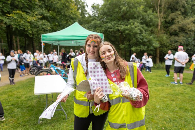 Waist-up shot of two teenage friends volunteering at an LGBTQIA pride colour run. They are both looking into the camera wearing high-visibility vests. The scene is filled with happiness, inclusivity, and community spirit, with people gathering in the background. They are located in Northumberland, England.

Videos are available similar to this Scenario.
