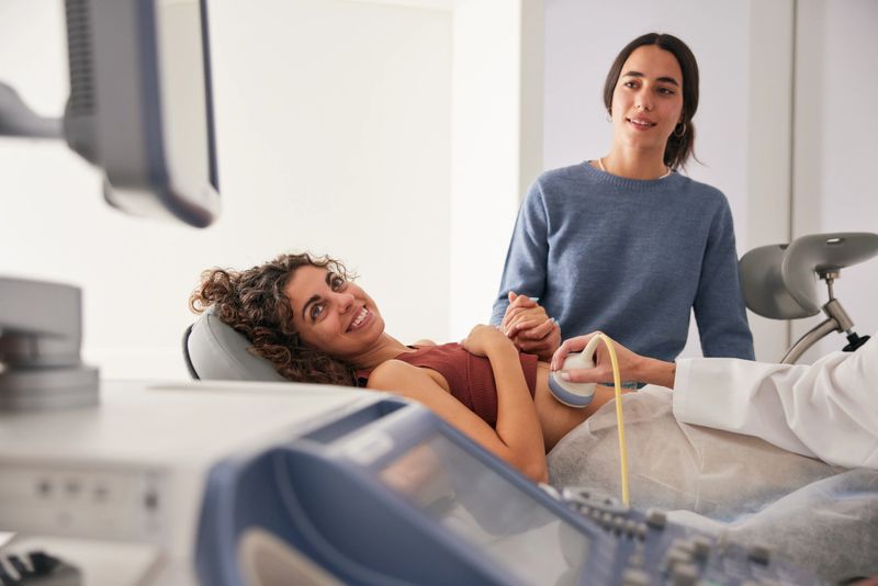 The young woman lies on the stretcher for an ultrasound, grasping her girlfriend's hand and looking at the scan screen.