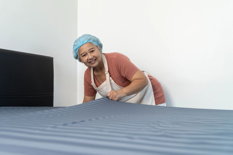 Smiling Middle aged housekeeper wearing cap and apron spreads the bedsheets in a bedroom, showcasing cleanliness and dedication to maintaining a tidy environment.