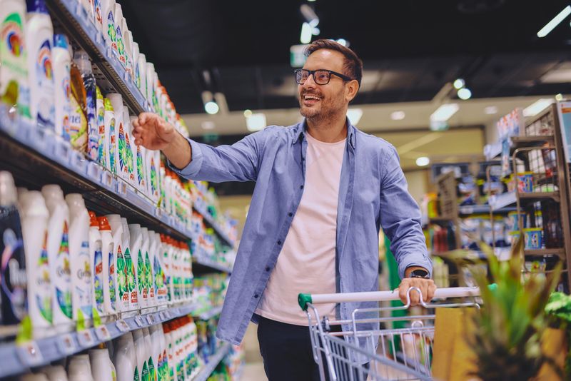 Adult man with bottle examining cleaning products on shelves while buying laundry detergent in supermarket