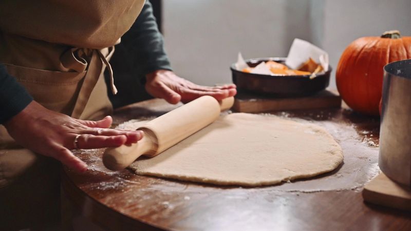 Baker's hands rolling dough on a wooden table, preparing a pumpkin pie. A rustic kitchen ambiance with natural light and the essence of homemade cuisine.