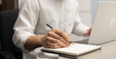 Person writing in a notebook while using a laptop.