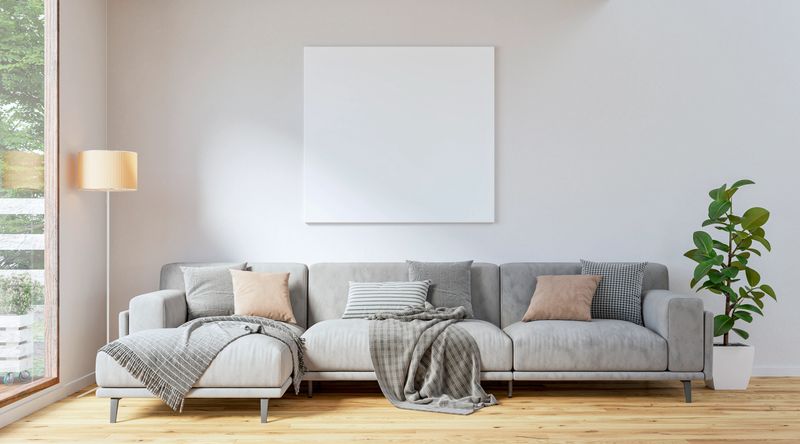 This modern living room features a sleek, neutral-toned sofa on natural oak hardwood floor, adorned with soft beige and grey cushions, along with cozy blankets. A large window brings in ample natural light, brightening the minimalist space. A potted plant (ficus) and a standing lamp add warmth, while the blank canvas on the wall invites creativity and personalization (copy space). 3D rendered image.