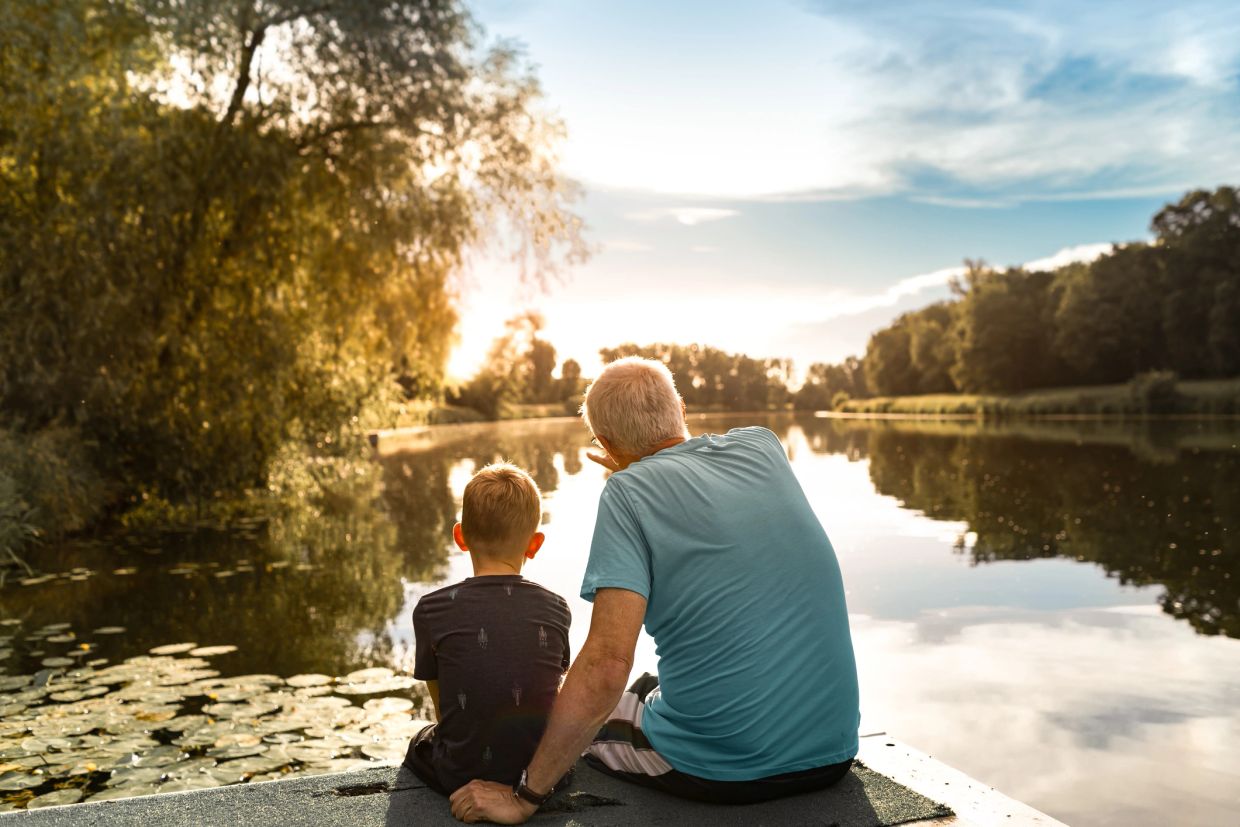 An elderly man and a boy sit by a lake at sunset.