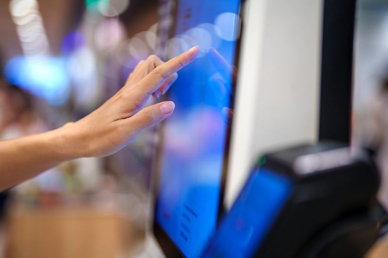The person is using their finger to confirm an order or payment for a product or service. The sleek, modern interface of the kiosk machine is illuminated, showcasing a simple, user-friendly layout.