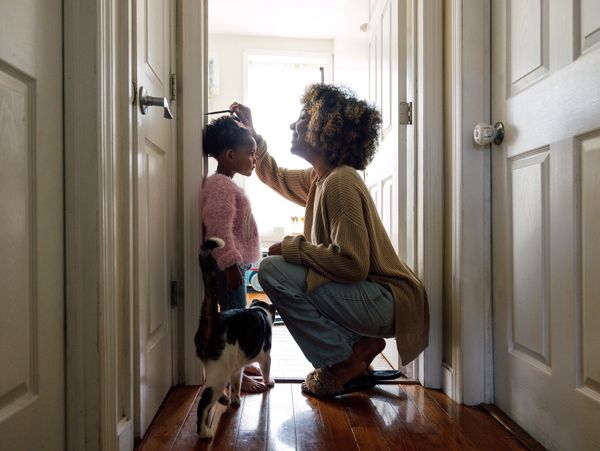 A woman combing a little girl's hair in a hallway while a cat walks by.