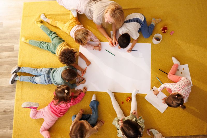 High angle view of a happy teacher and little children drawing on big white paper together in the kindergarten