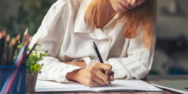 Woman sat at a desk holding a pen and writing in a note book.