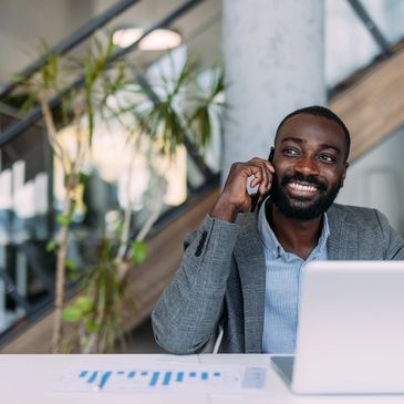 Smiling man in a suit talking on phone at his desk with laptop and chart.