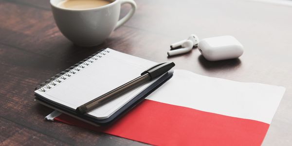 A cup of coffee, notebook, pen, and wireless earbuds on a wooden table with a red and white cloth.