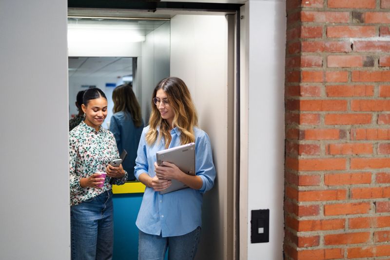 Two business women are exiting an elevator together while using a smartphone and a tablet