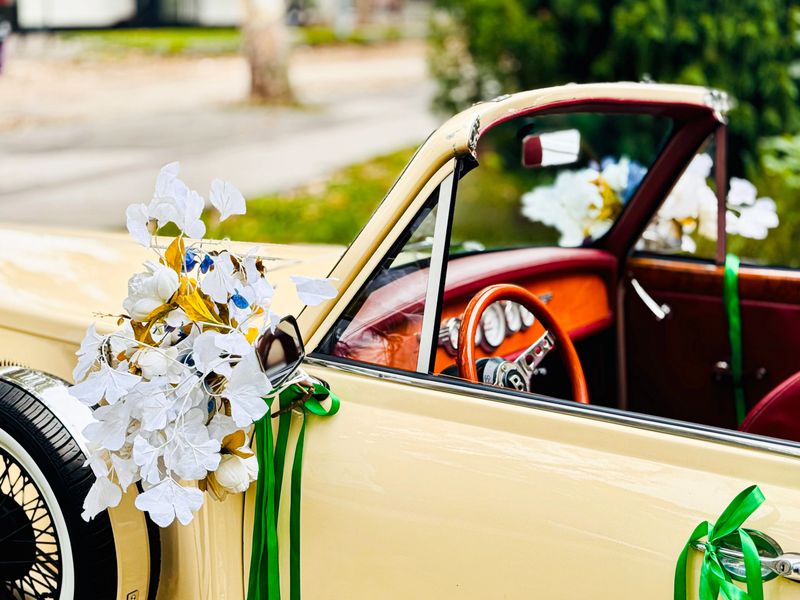 A classic beige convertible is adorned with white floral arrangements and green ribbons for a wedding event, parked in a scenic outdoor setting.