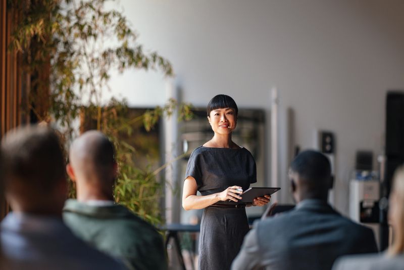 A confident presenter leads a business conference, engaging a diverse audience. The speaker is using a tablet and microphone, ensuring effective communication and interaction in a modern professional setting.