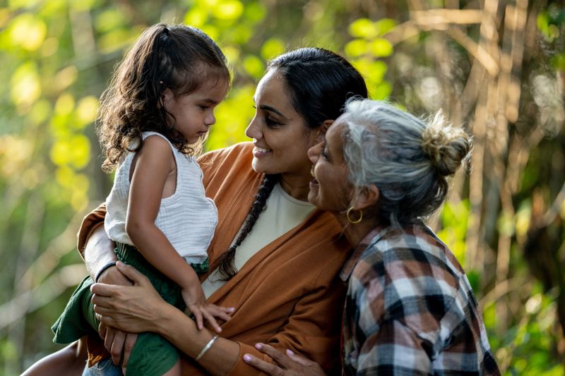 Three generations of women pose together outside for a portrait.  A Mother holds her daughter in her arms as the Grandmother leans in closely and they share a laugh.