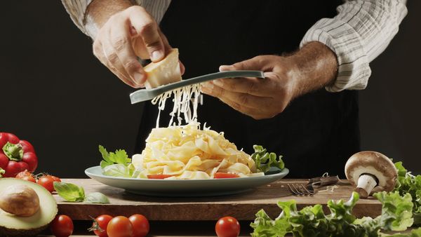 Chef grating cheese over pasta surrounded by fresh vegetables.