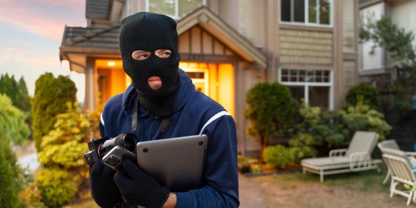 A masked burglar holding a camera and tablet outside a house at dusk.