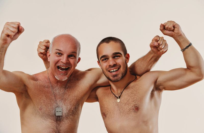 Two athletic men are flexing their muscles and smiling, embodying the joy of physical power and friendship. They are both wearing necklaces, highlighting their bond