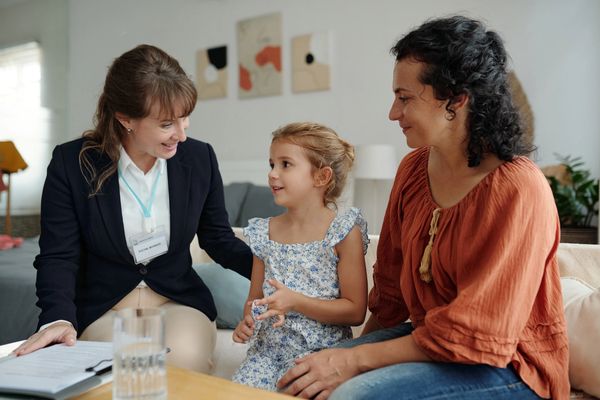 
A professional woman in a dark blazer is sitting on a couch with a mother and her young daughter.