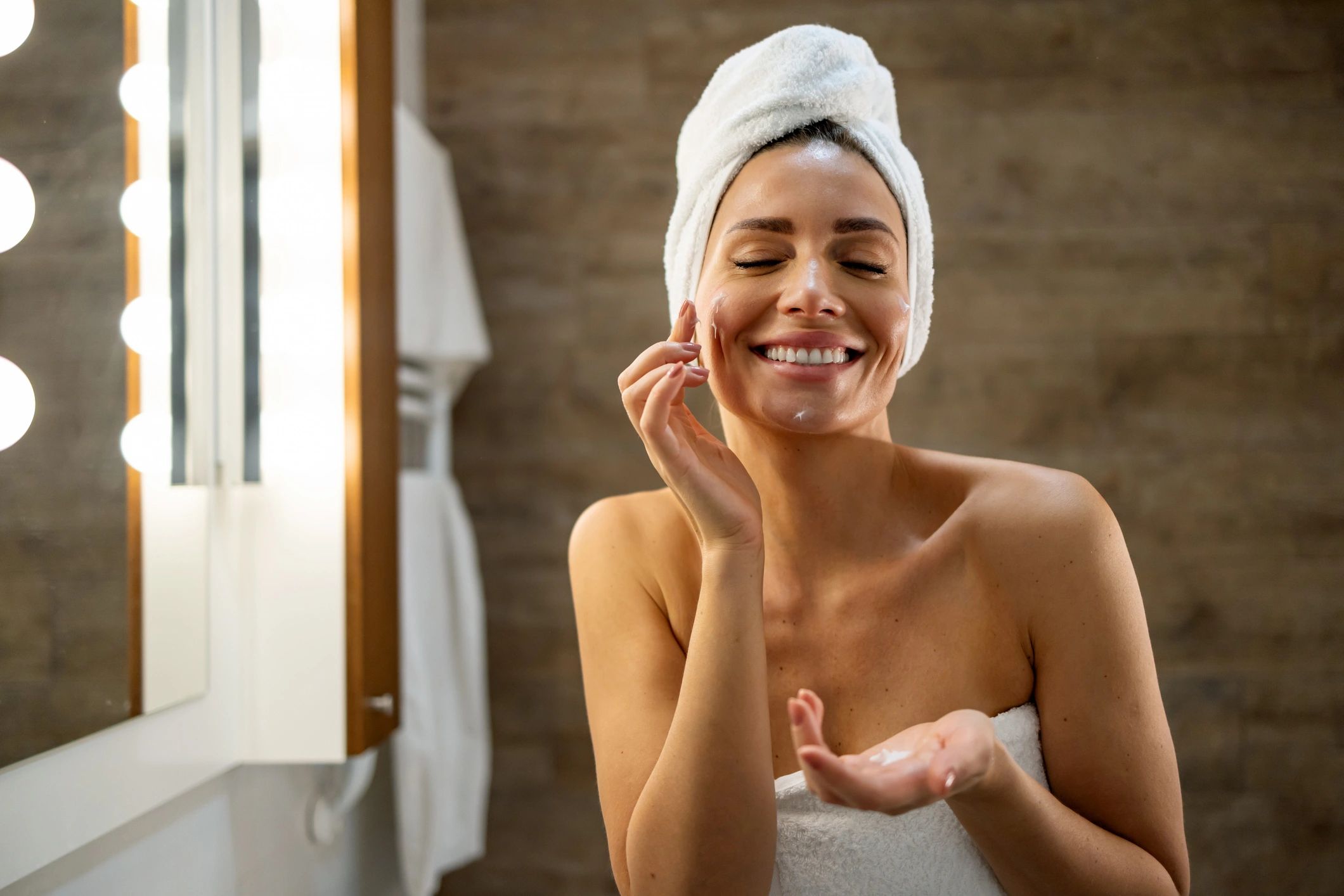 Woman with towel on head applying face cream in bathroom mirror.