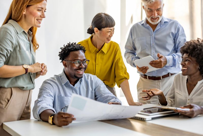A group of diverse professionals engaged in a positive meeting, sharing ideas and documents in a bright office setting. The atmosphere is collaborative and inclusive.