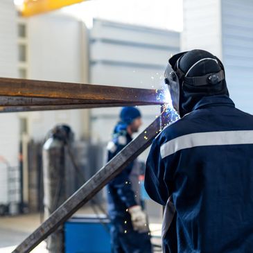 Welder wearing protective gear sparks while welding metal beams indoors.