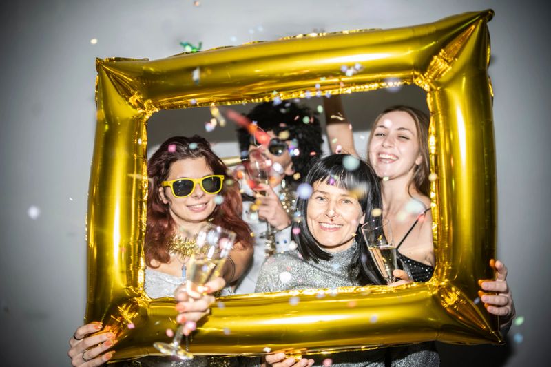 Four friends hold a golden balloon frame, celebrating with champagne and confetti, radiating joy and festive spirit at a party. Vibrant and lively atmosphere captured.