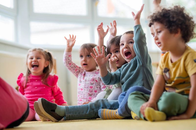happy little children are sitting in the kindergarten on the floor with raised arms, laughing, happy, playing