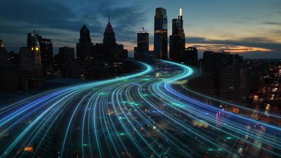 City skyline with glowing blue light trails on a highway at dusk.