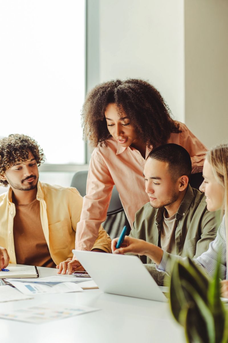 Diverse team of young professionals are collaborating on a project, using a laptop and going over paperwork in a bright office