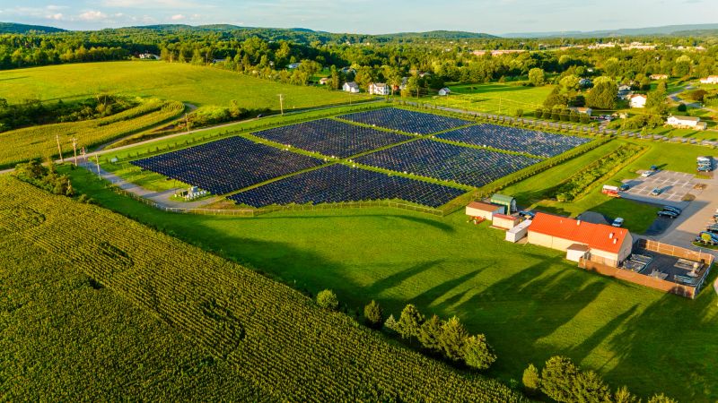 Expansive solar panel array situated in rural landscape in Pennsylvania. Lush green fields, farmland, and forested hills surround the installation. Scattered houses and buildings of a small community dot the vicinity. The orderly arrangement of dark solar panels contrasts with organic patterns of surrounding agricultural fields and natural vegetation.