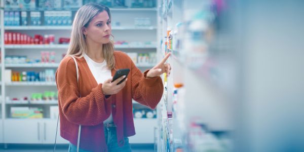 A woman in a pharmacy selecting medication while holding her phone.