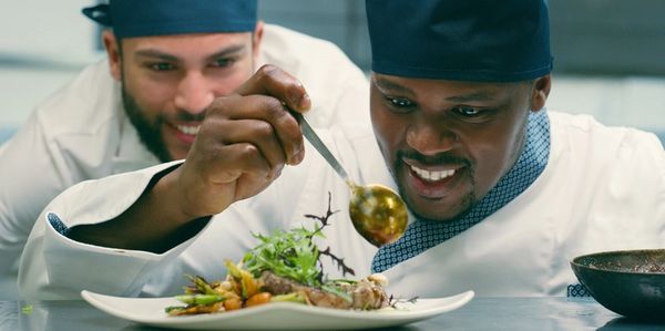 Two chefs carefully plating a gourmet dish with sauce and greens.
