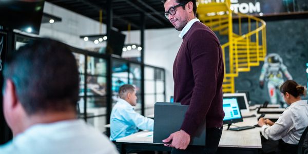 A man holding a laptop stands in a modern office with colleagues working at desks.
