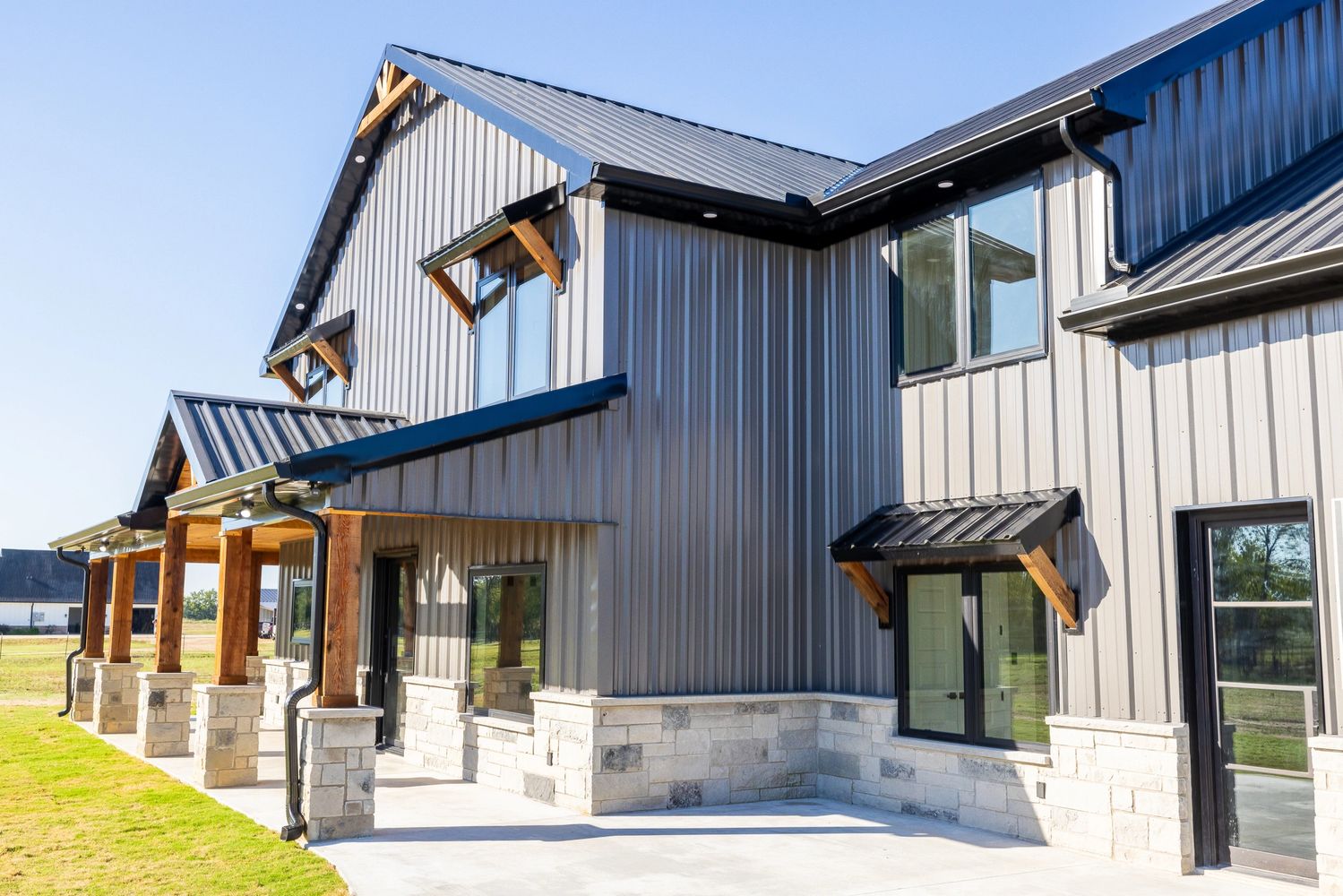 Modern two-story metal house with stone accents and wooden pillars under a clear blue sky.