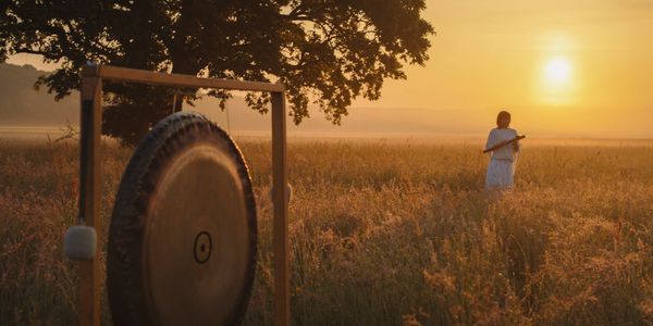 A person in white stands in a field at sunset near a large gong.