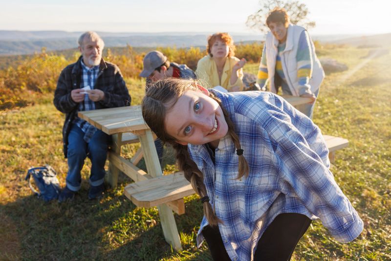 Cheerful young girl in blue plaid shirt leans towards camera, smiling brightly. Family members gather around wooden picnic table on grassy hilltop. Scenic Poconos landscape with rolling hills stretches in background. Warm golden light of late afternoon creates cozy atmosphere for family outing.
