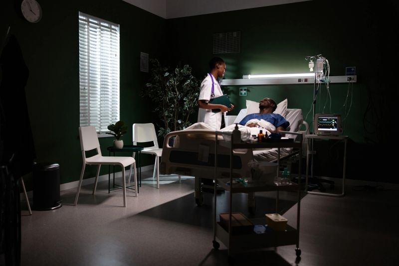 Simple moment in the hospital room, an African girl stands with a folder beside a patient lying in bed.