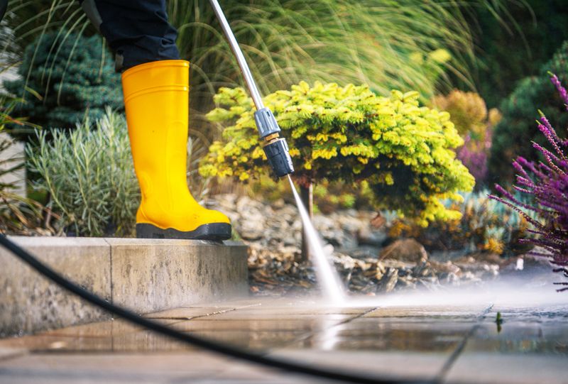 A person is using a pressure washer to clean a patio surface while standing in a beautifully landscaped garden. The vibrant greenery and flowers create an inviting atmosphere.
