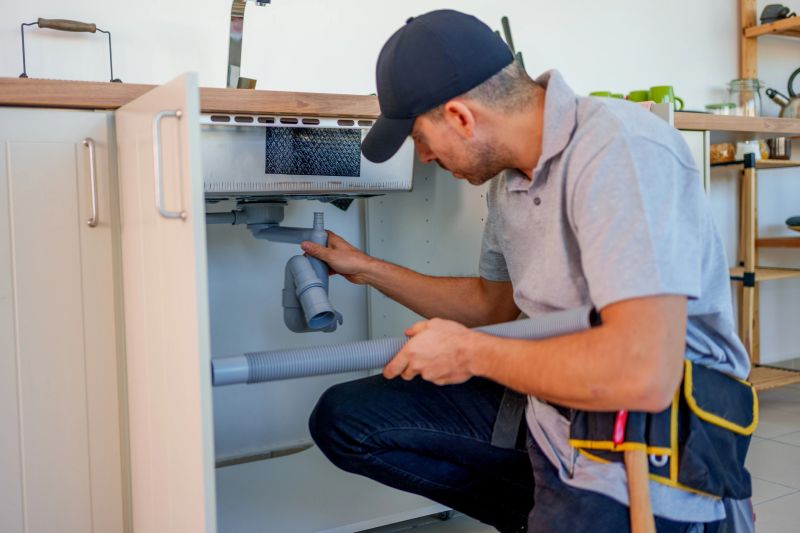This image depicts a handyman or technician working on the plumbing under a kitchen sink. He is wearing a grey polo shirt and a black cap, and  to be inspecting and installing grey plastic pipes. The cabinet door beneath the sink is open, and the man is focused on fitting or securing the plumbing system. He is holding a piece of tubing, preparing to connect it. The setting seems to be a home kitchen, and the person is clearly engaged in a repair or installation task involving the sink's drainage system. The tool belt he is wearing suggests he is equipped for various tasks.