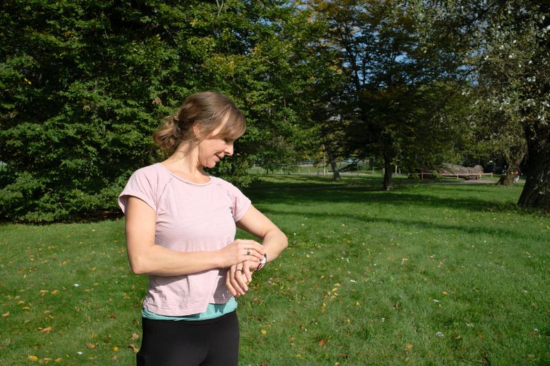A woman checks her fitness tracker during a morning exercise in the public park, ensuring she stays on track with her goals. She appears focused and content, appreciating the balance between technology and nature. The calm waters and lush greenery around her reflect a moment of mindfulness and dedication to self-care.