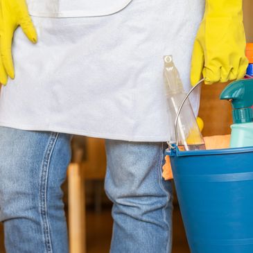 Person holding a blue bucket with cleaning supplies, wearing yellow gloves and jeans.